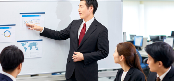 Man in a suit pointing at a whiteboard with a group of people listening in an office setting.
