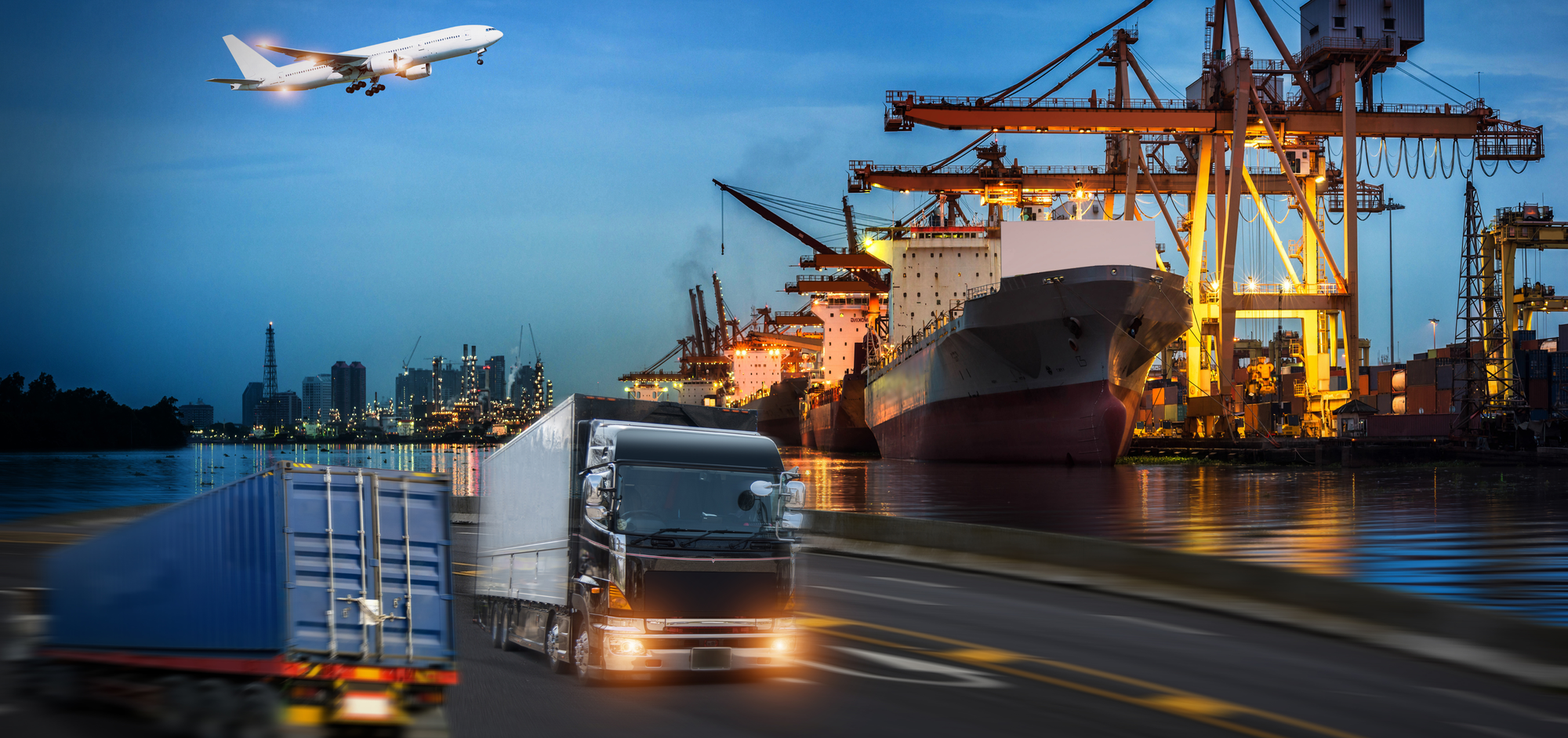 Truck driving on a road with a ship and airplane in an industrial harbor at night.